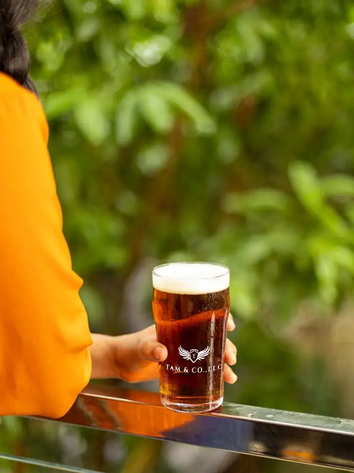 A hand holding a pint of Tam & Co. beer against green foliage